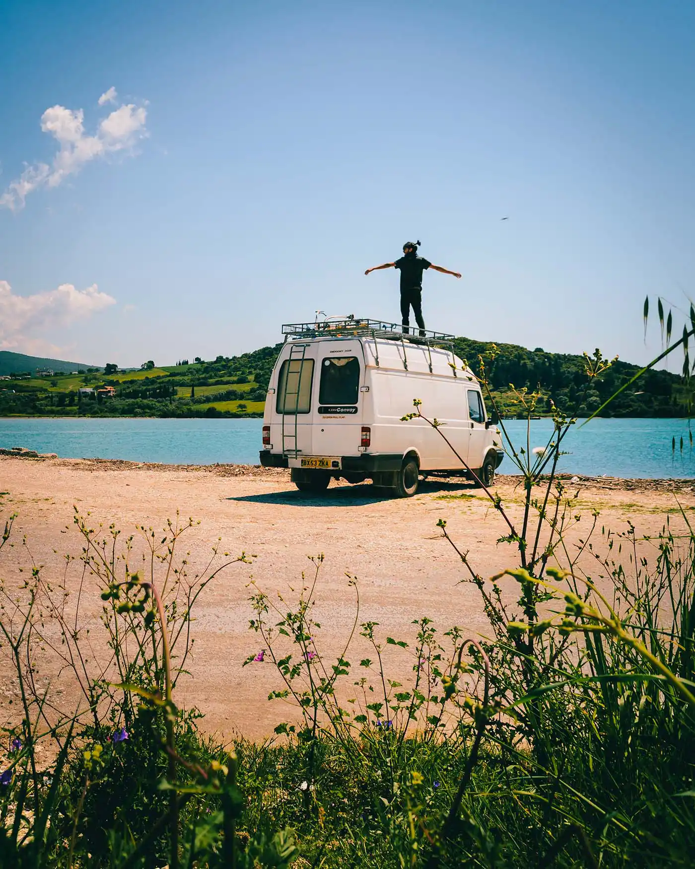 The van parked above a Mediterranean coastline, blue water below, sun out.